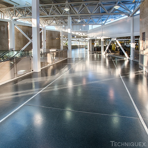 Bright, spacious interior walkway with polished dark flooring, metal railings, and exposed beams, featuring escalators and large windows letting in natural light.