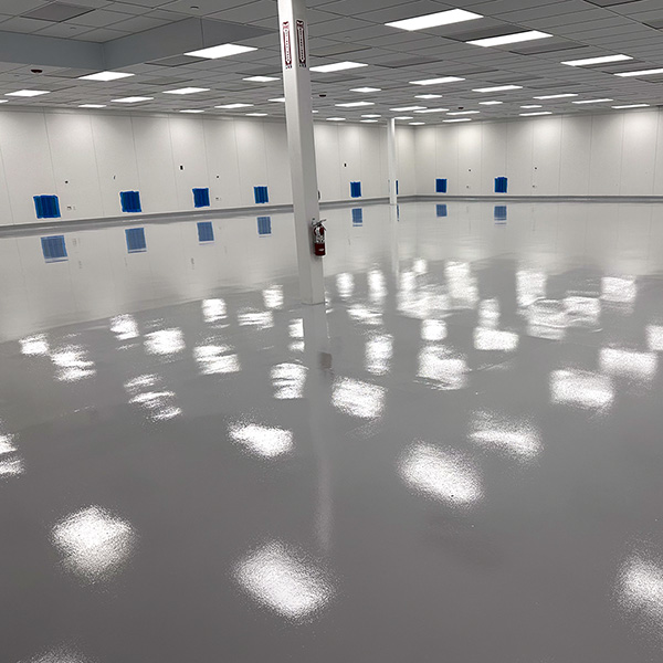 Large empty room with glossy, reflective flooring, white walls, a grid ceiling with bright lights, and a fire extinguisher mounted on a column.