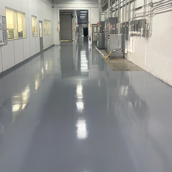 A clean, shiny gray floor in an industrial hallway with electrical panels and windows on the left wall.
