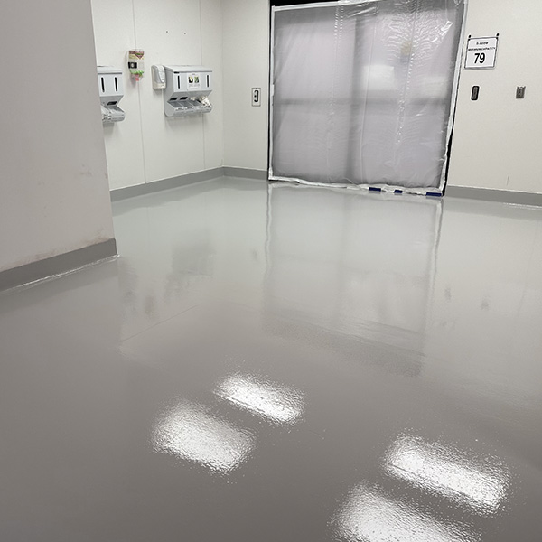 A clean, empty room with glossy, light gray epoxy flooring, white walls, wall-mounted dispensers, and a plastic-covered window or door at the back.