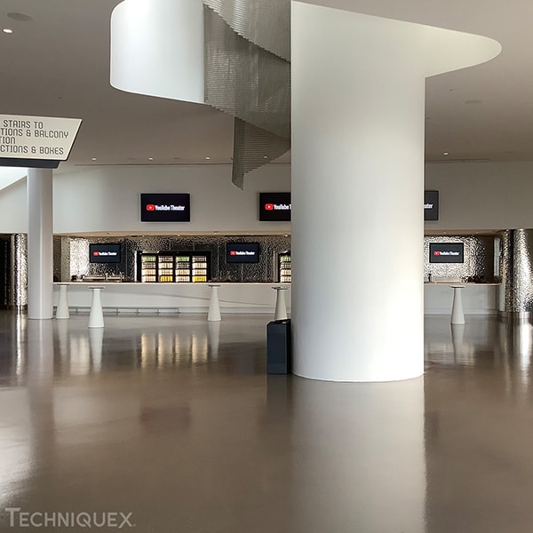Modern lobby area with a large white column, digital screens, and a sign indicating directions to stairs, restrooms, and balcony. The floor is reflective and the space appears empty.