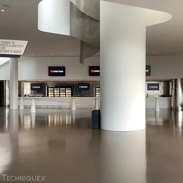 Modern lobby area with a large white column, digital screens, and a sign indicating directions to stairs, restrooms, and balcony. The floor is reflective and the space appears empty.