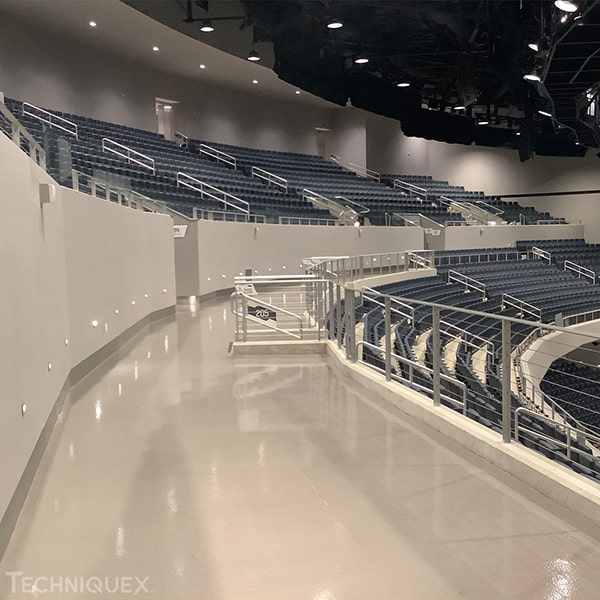 Indoor arena with empty dark blue stadium seats, a curved walkway with metal railings, and overhead lighting. The area is well-lit and appears clean and modern.