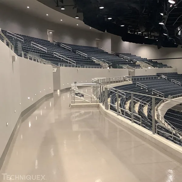 Indoor arena with empty dark blue stadium seats, a curved walkway with metal railings, and overhead lighting. The area is well-lit and appears clean and modern.