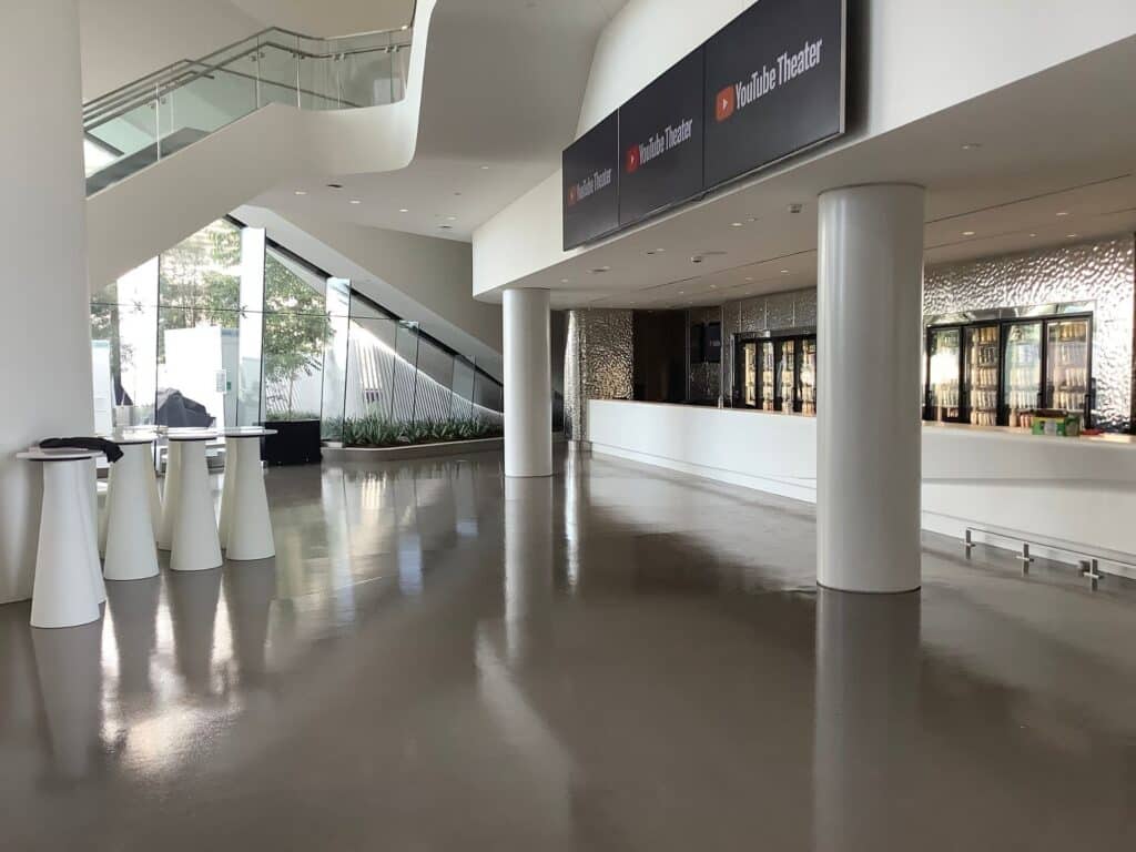 Spacious, modern lobby with shiny floors, a staircase, tall tables, large windows, and a concession counter labeled "YouTube Theater.