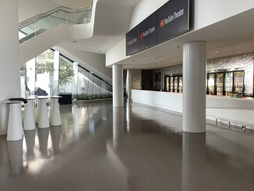 Spacious, modern lobby with shiny floors, a staircase, tall tables, large windows, and a concession counter labeled "YouTube Theater.