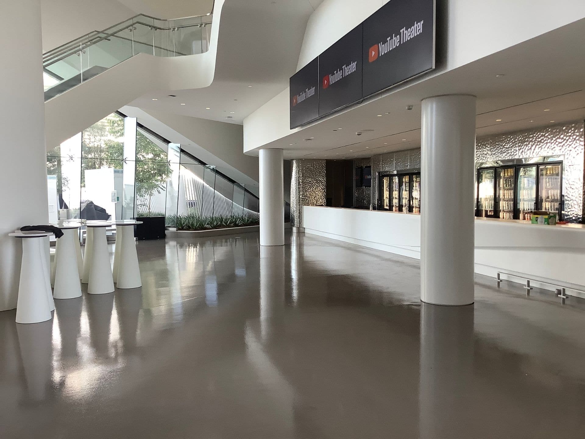 Spacious, modern lobby with shiny floors, a staircase, tall tables, large windows, and a concession counter labeled "YouTube Theater.