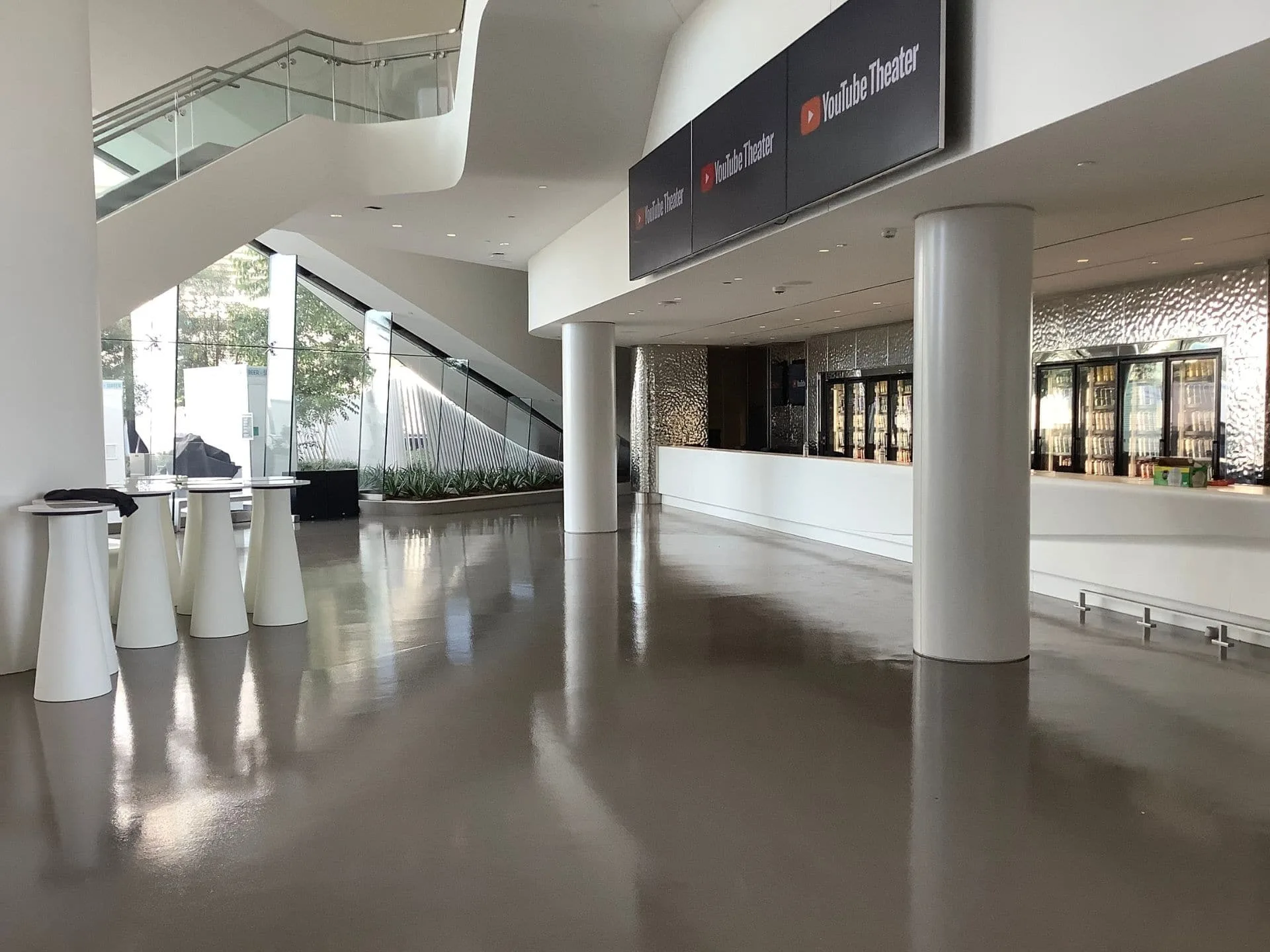 Spacious, modern lobby with shiny floors, a staircase, tall tables, large windows, and a concession counter labeled "YouTube Theater.