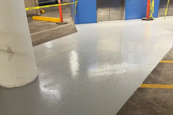 A freshly applied gray epoxy floor in a parking garage, cordoned off by yellow caution tape and traffic cones in front of an elevator.