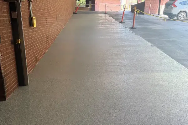 A smooth, freshly paved gray concrete walkway along a brick building wall, leading toward a parking area with safety cones and yellow caution tape.