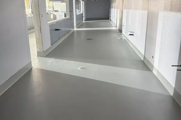 A wide, sterile hallway under construction with gray flooring and white walls, featuring an open service window and an exposed ceiling structure.